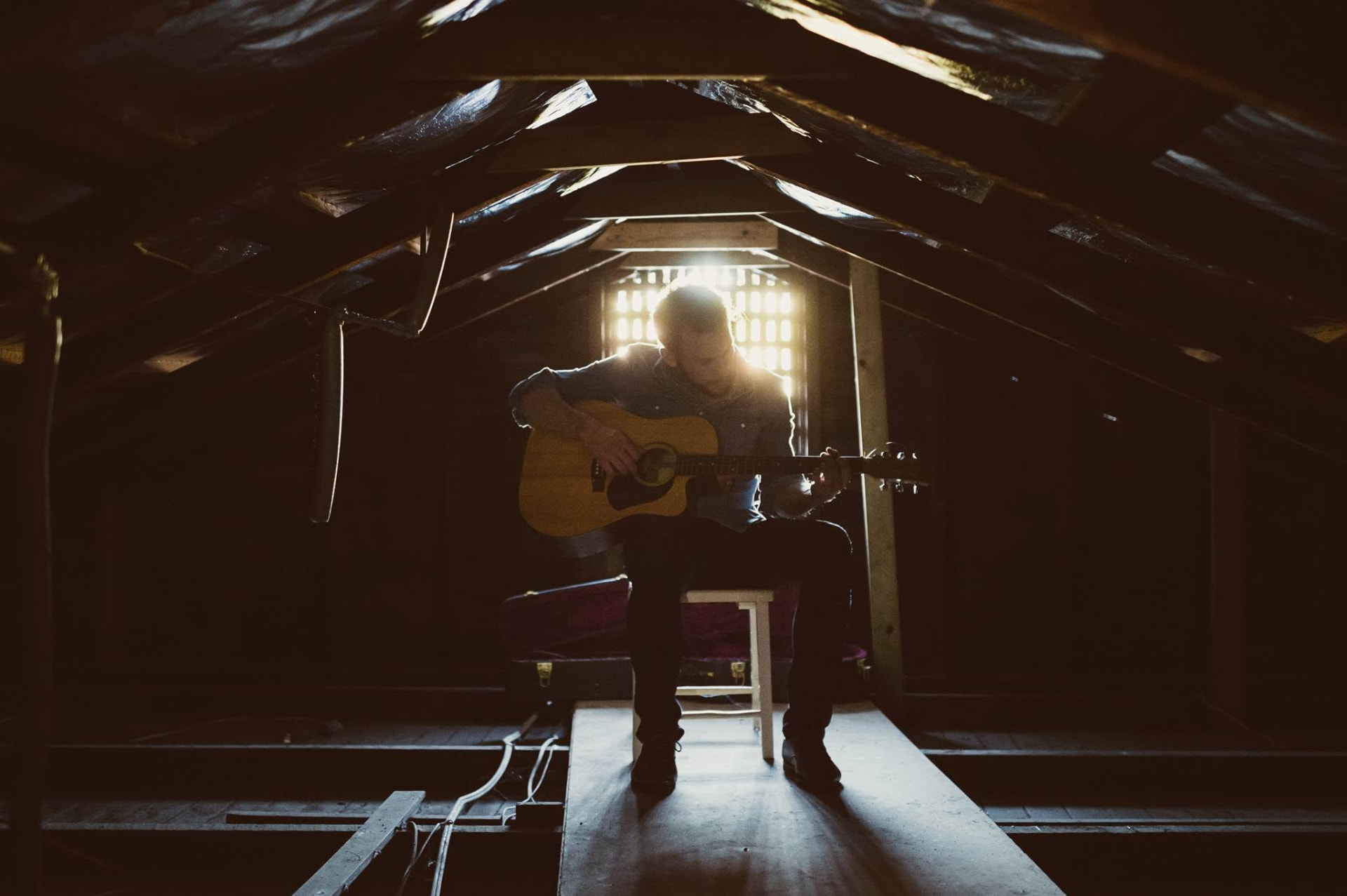 Dan Smith playing acoustic guitar in a moody backlit attic, light streaming through a window behind him